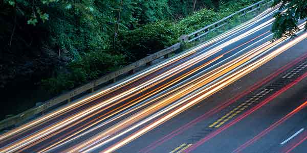 Long exposure of car light trails on a highway, green foliage on the side.