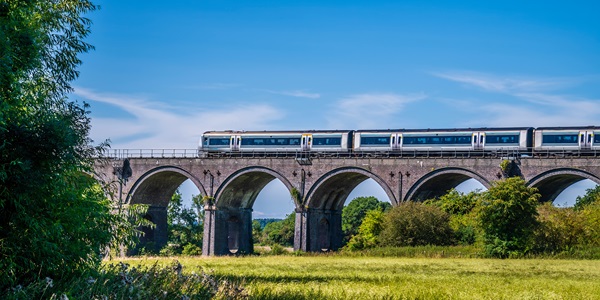 Train crossing a stone viaduct over grassy field under clear blue sky.